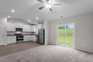 A modern, open-concept living space features a kitchen with white cabinets, stainless steel appliances, and a wood floor. The carpeted living area has a ceiling fan and large sliding glass doors leading to a grassy yard.