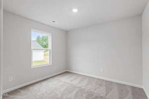 An empty room with light gray walls and beige carpet. There's a single window on the left wall allowing natural light inside. A ceiling light provides additional illumination.