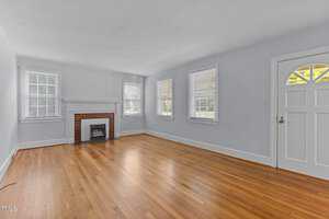 An empty living room with light blue walls, hardwood floors, a white fireplace with a brick interior, three windows with white blinds, and a white front door with a semi-circular window design. Natural light illuminates the space.
