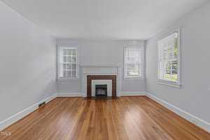 An empty room with light blue walls, hardwood floors, and a brick fireplace centered on one wall. Two windows with white blinds flank the fireplace, letting in natural light. The ceiling is white, and the overall atmosphere is bright and airy.
