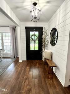 A foyer with a black front door featuring a window and wreath. There is a wooden bench with a cushion to the right and a round mirror above it. The floor is wooden, and natural light streams in through a nearby window.