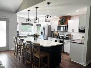 Modern kitchen with a large island, wooden stools, and pendant lights. Stainless steel appliances and white cabinets enhance the sleek design. A plant decorates the wall, and natural light fills the room through large windows.