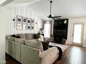 A cozy living room with a large beige sectional sofa, a wooden coffee table, and a patterned rug. The walls are decorated with framed photos. There's a fireplace with a mantel, a ceiling fan, and sheer curtains on the windows. A door leads outside.