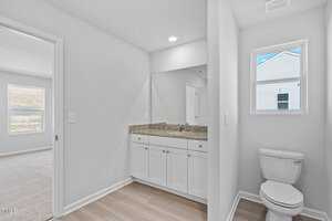 A bright bathroom with white walls and light wood flooring. It features a white cabinet with a granite countertop, a large mirror, and a toilet. A window lets in natural light. Carpeted room visible to the left.