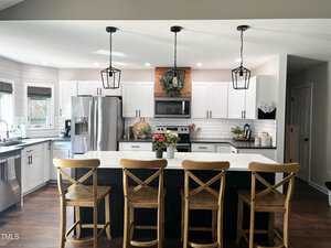 Modern kitchen with a large island featuring four wooden chairs. White cabinets and stainless steel appliances are present, along with pendant lights overhead. A decorative wreath is above the stove, adding a touch of green.