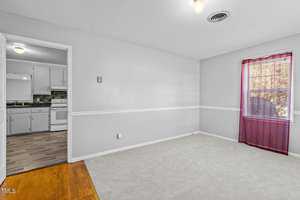 A small, empty room with light gray walls, white trim, and a tan carpet. A window with sheer red curtains is on the right. An open doorway leads to a kitchen with white cabinets and an electric stove. A ceiling light fixture is visible.