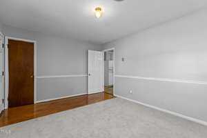 An empty room with light gray walls, hardwood flooring, and a white carpeted area. A single ceiling light is turned on. There are two doors, one closed with dark wood and one open leading to another space.