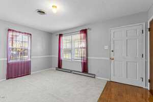 An empty room with light gray walls, white trim, and a mix of carpet and hardwood flooring. It has two windows with red curtains and a white door. A single ceiling light is on.