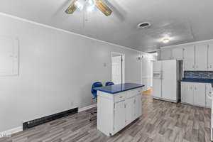A kitchen with a white fridge, blue countertop island with two blue chairs, and white cabinets. The floor features gray patterned tiles, and a ceiling fan with wooden blades is visible. Walls are a light gray color.