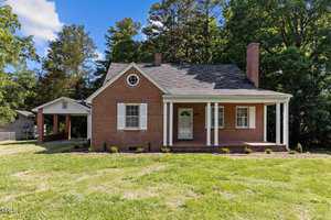 A brick house with a covered porch and two white columns, set against a backdrop of tall trees. It has a gabled roof, white shutters, and a small, manicured lawn in the foreground. A carport is visible on the left side.