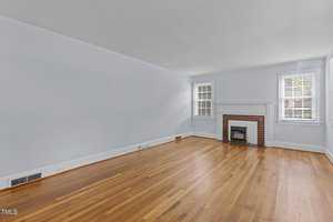 A bright empty room with light blue walls and wooden floors. It features a white-trimmed fireplace with a brick surround. Two windows with white shutters let in natural light.