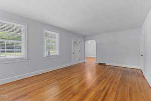 A bright, empty living room with light blue walls, hardwood floors, and two windows with white blinds. A doorway leads to another room with similar flooring. Natural light fills the space, creating a welcoming atmosphere.