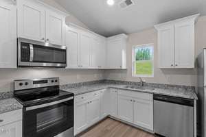 A modern kitchen with white cabinets and granite countertops. It features a stainless steel oven, microwave, and dishwasher. A small window above the sink provides natural light, and the room has a wooden floor.