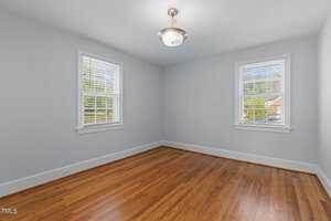 A simple, empty room with light blue walls and hardwood floors. Two windows with white blinds allow natural light to enter. A ceiling light fixture is centered above.