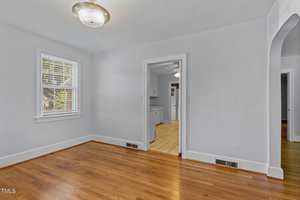 A bright, empty room with hardwood floors and light blue walls. There's a ceiling light fixture and a window with blinds on the left. The doorway leads to a kitchen area with white cabinets and wooden flooring.