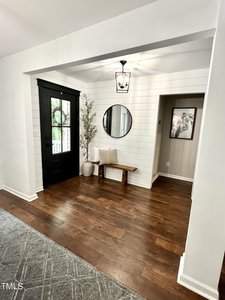 A modern entryway featuring a black door with a glass panel, a wooden bench beneath a round mirror on shiplap walls, and a small tree in a vase. A black lantern-style light fixture hangs from the ceiling. The floor is dark wood with a gray carpet nearby.