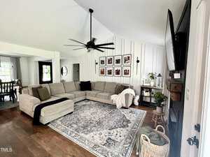 A spacious living room with a large beige sectional sofa, a patterned area rug, and a ceiling fan. A flat-screen TV is mounted on the wall, and family photos are arranged in a grid on another wall. Plants and wicker baskets add cozy accents.