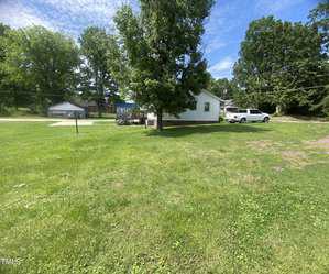 A grassy yard with a large tree in the center. A white house and a white car are visible in the background, surrounded by green trees. The sky is clear with a few clouds.