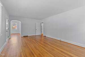 An empty room with light blue walls and hardwood flooring. There is an open doorway leading to another room and a double window on the left. A white door and an archway are visible in the background. The room is well-lit with natural light.