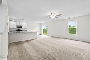 Spacious empty room with beige carpet, white walls, and a ceiling fan. There's a kitchen with white cabinets and stainless steel appliances on the left, and large windows offering a view of greenery on the right.