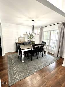 A dining room with a rectangular wooden table and six chairs. A chandelier hangs above. A side table holds decor and plants near a large window with light curtains. The room has white walls, hardwood floors, and a patterned gray rug.