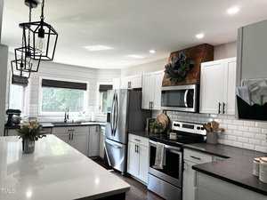 Modern kitchen with white cabinets and dark countertops. Stainless steel appliances include a fridge and an oven with a microwave above. A large island features hanging geometric light fixtures. A window allows natural light, and greenery decorates the space.