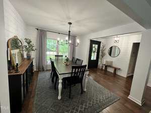 A dining room with a large white table, surrounded by black chairs. A decorative rug covers the wooden floor. A sideboard with artwork and plants is against the wall. A window with curtains lets in natural light. A door leads to another room.