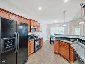 A spacious kitchen with wooden cabinets, black appliances, and a tiled floor. The room features a double-door fridge, oven, microwave, and a sink on a central island. A dining area with a table and chairs is visible in the background. Natural light streams in.