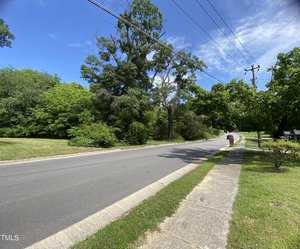 A quiet suburban street lined with green trees under a clear blue sky. A sidewalk runs alongside the road, and a person walks in the distance. Power lines stretch overhead, and the area is lush with vegetation.
