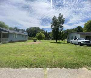 A grassy yard stretches between two single-story houses under a partly cloudy sky at 815 Goshen Street, Oxford. A white vehicle is parked in the driveway of the house on the right, and trees line the back of the property.