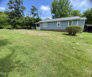 A light blue house with a metal roof sits on a large grassy yard dotted with small pink flowers. The porch is white, and there are trees and bushes surrounding the property. The sky is clear and sunny.