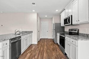 A modern kitchen with white cabinets and gray granite countertops. It features stainless steel appliances including a refrigerator, oven, and dishwasher. The floor is wooden, and there are recessed lights on the ceiling.