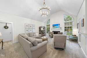 A cozy living room with vaulted ceilings, featuring a large sectional sofa and two armchairs facing a fireplace. A chandelier hangs above, and a flat-screen TV is mounted above the mantel. Large arched windows bring in natural light and greenery views.