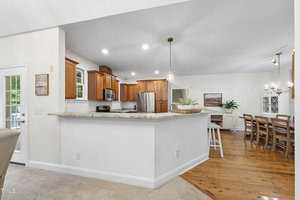 A modern kitchen with wooden cabinets and stainless steel appliances, including a refrigerator and oven. An island with a marble countertop and bar stools separates the kitchen from the spacious dining area with a long wooden table and chairs.