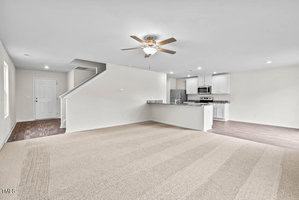 Spacious empty living room with beige carpet, adjoining a kitchen area with stainless steel appliances and white cabinets. Ceiling fan and neutral walls, with a staircase and door leading off the space.