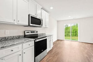 A modern kitchen with white cabinets, granite countertops, and stainless steel appliances, including a stove and microwave. The room has wooden flooring and large sliding glass doors leading to a garden with lush green trees.