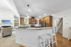 A modern kitchen with white walls, wooden cabinets, and a marble-topped island featuring two white bar stools. Stainless steel appliances are present, and a cozy living area with a fireplace and armchair is visible in the background.