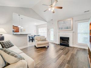 Spacious living room with wooden floors, beige sofa, armchair, and a fireplace with art above. Ceiling fan overhead, large windows, and doorway to an adjacent dining area with a table and chairs in the background.