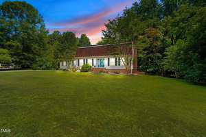 A white house with a brown roof is surrounded by a manicured lawn and lush trees. The sky is a mix of blue, pink, and orange hues, indicating sunset. A turquoise door and small shrubs line the front of the house.