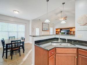 Bright, modern kitchen and dining area with dark granite countertops, wooden cabinets, and a double sink. A wooden dining table is set for six near large windows, and a ceiling fan and wall decor are visible in the background.