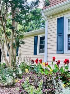 A charming house with yellow siding and blue shutters is surrounded by a lush garden. Red flowers, green foliage, and tall trees are in the foreground, creating a vibrant and inviting atmosphere.