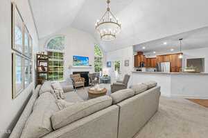 Spacious living room with a large gray sectional sofa, modern chandelier, and a TV above the fireplace. Large windows let in natural light, and a view of the kitchen with wooden cabinets is visible. Neutral decor with a cozy, inviting atmosphere.