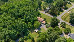 Aerial view of a suburban area surrounded by dense green trees, featuring a house with a driveway and a smaller outbuilding nearby. Roads border the property, leading into a neighborhood with similar homes.