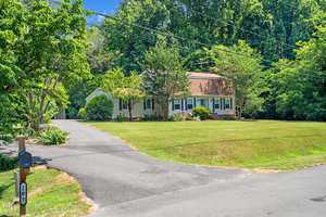A charming suburban house with a brown roof and light-colored siding is surrounded by a well-maintained lawn and lush trees. A paved driveway leads to the house, and a mailbox labeled "20" is visible in the foreground.