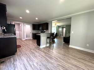 Spacious open-plan kitchen and living area with light wood flooring. The room features dark wood cabinets, a kitchen island with bar stools, and a view into a well-lit living room. A laundry area is visible to the left.