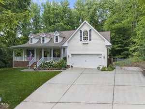 A suburban white house with a gabled roof and attached garage sits surrounded by lush greenery. A well-maintained lawn and a small garden border the driveway. An American flag hangs by the front porch.