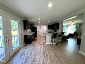 Open-concept kitchen and living area with wood flooring. The kitchen features dark cabinets, stainless steel appliances, and a breakfast bar with stools. French doors on the left lead to a patio. The living room has a large window and ceiling fan.