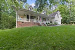 A charming white house with a brick facade, featuring a covered porch adorned with hanging plants and an American flag. It is set amidst lush greenery with a well-manicured lawn and surrounded by tall trees under a clear sky.