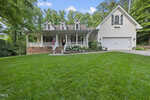 At 568 Timberknoll Lane in Wake Forest, this charming two-story house boasts a white exterior and a front porch with columns adorned by an American flag. The property features a well-manicured lawn, a flower bed, and a driveway leading to a garage, all surrounded by tall green trees under the blue sky.