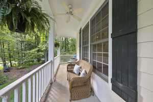 A cozy porch with wicker furniture, including a loveseat with cushions, is adorned with a hanging fern. The porch is surrounded by a white railing and faces a green, wooded view. A ceiling fan hangs above for comfort.